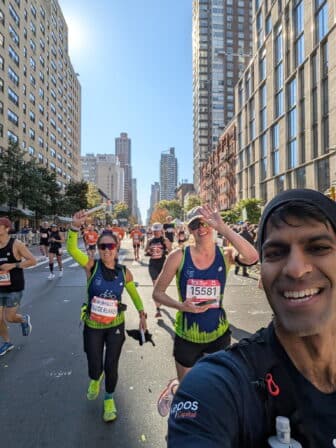 Runners participate in a city marathon. A man in the foreground takes a selfie, smiling. Behind him, two women in race bibs wave at the camera. Tall buildings line both sides of the sunny street.