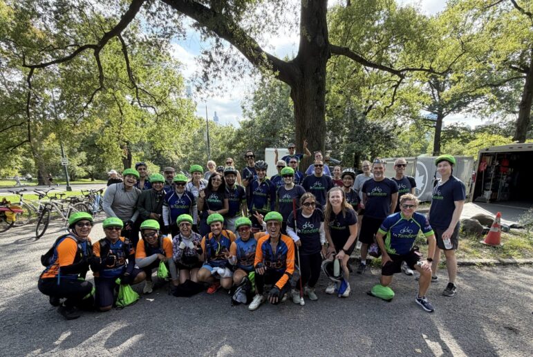 A large group of cyclists wearing green helmets and athletic gear pose for a group photo outdoors on a sunny day with trees and bicycles in the background.