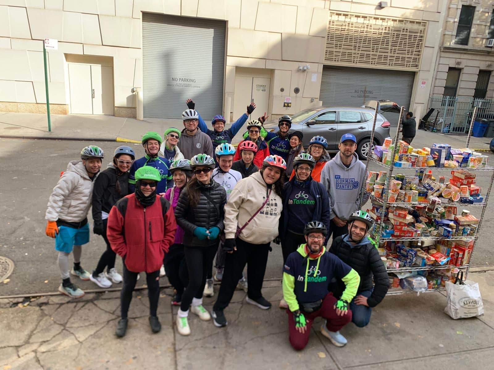 A group of cyclists wearing helmets stands with a metal cart overflowing with canned food and boxed groceries on a city sidewalk.