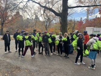 A group of people wearing bike helmets and carrying bright green In Tandem backpacks gather in a park.