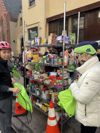 Two cyclists in helmets fill bright green bags with groceries from a multi-tiered wire shelving unit stocked with canned and boxed food outside a pantry.